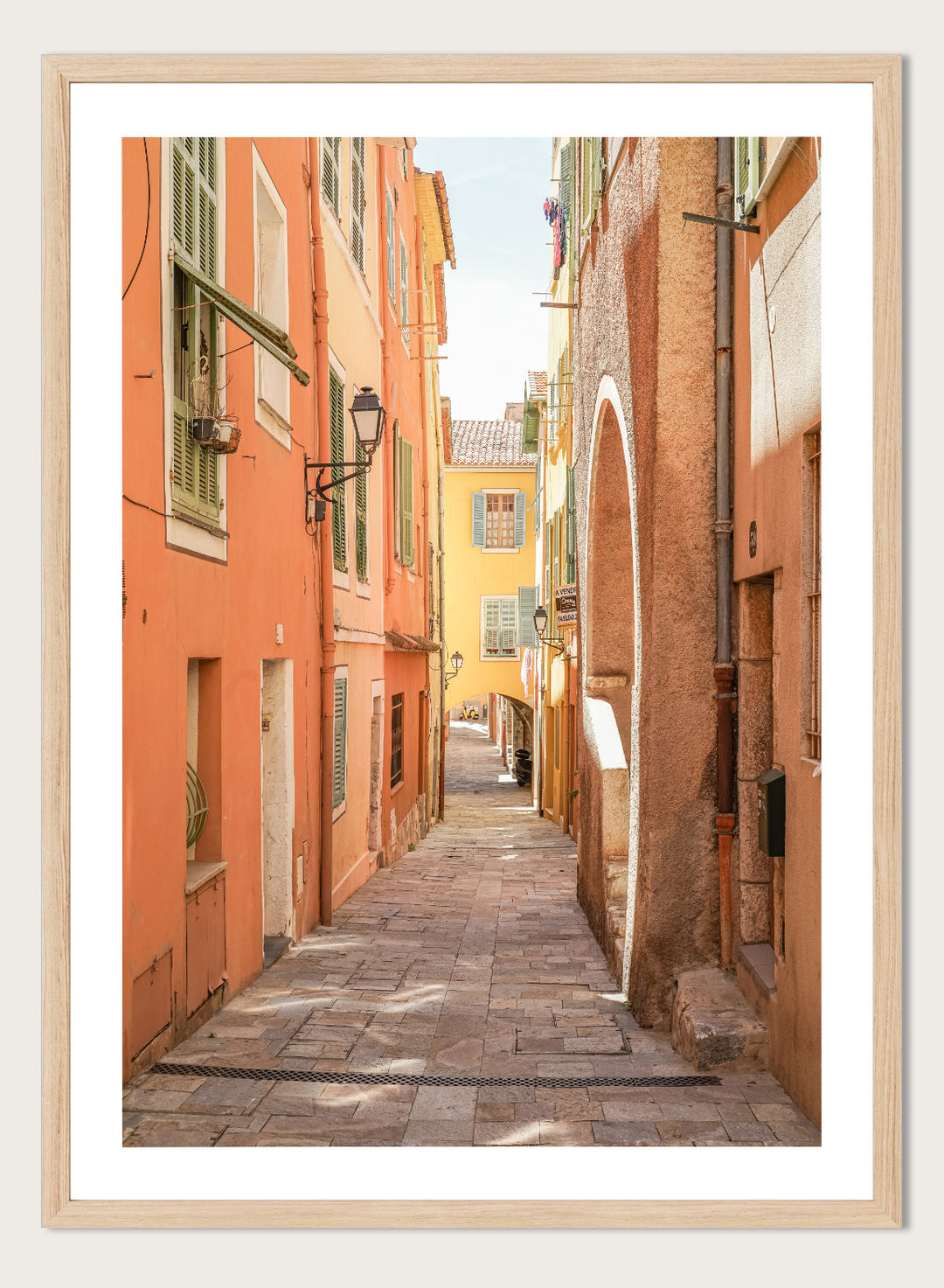 Framed photograph of a narrow street on a wall next to a window.  narrow street with colorful buildings in a European city. Vibrant European Alley by Henrike Schenk is a travel photography print featuring a Mediterranean street, cobblestone alley, and pastel European architecture.