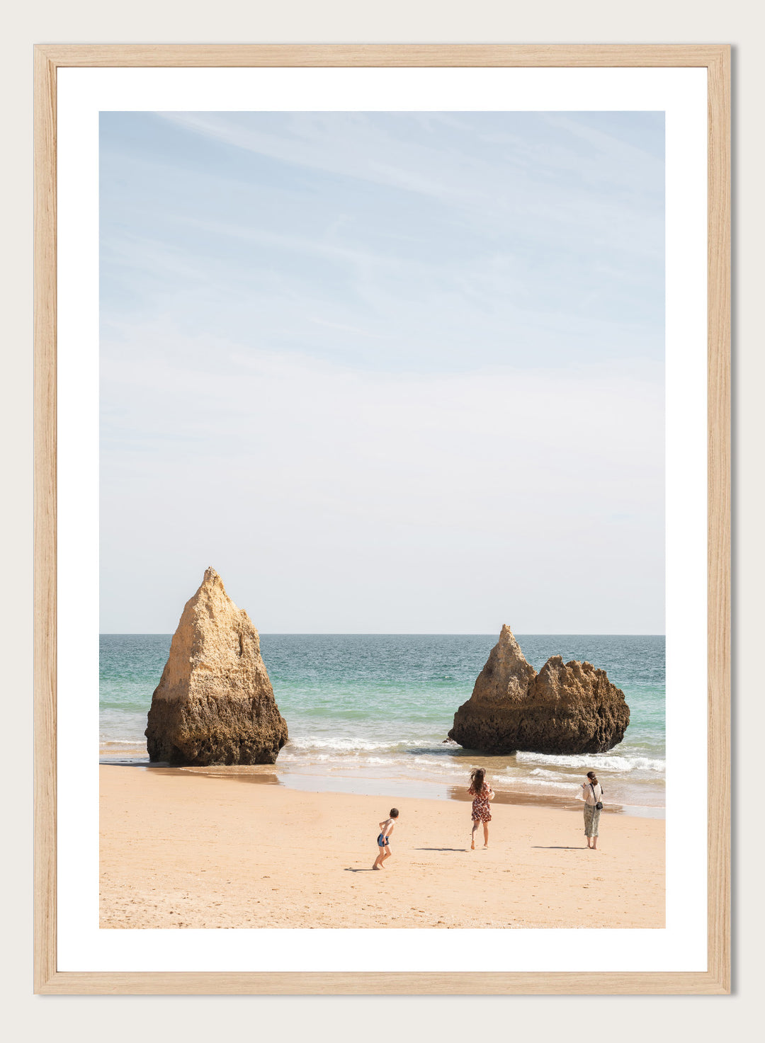 Three people on a beach with large rock formations and clear blue sky. Family Beach Day by Henrike Schenk is a coastal photography print showing a family on a sandy beach with ocean waves and dramatic sea cliffs.