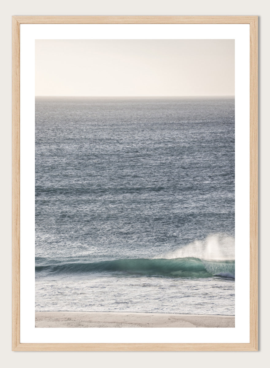 A framed photographic art print of an ocean scene, featuring a gentle wave breaking on a sandy beach under a pale sky. Tranquil Ocean Scene by Shot by Clint ocean photography print with a rolling wave, soft sea spray, and airy blue coastal tones.