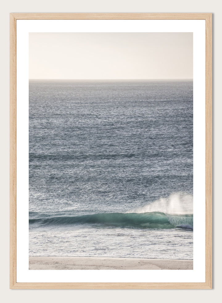 A framed photographic art print of an ocean scene, featuring a gentle wave breaking on a sandy beach under a pale sky. Tranquil Ocean Scene by Shot by Clint ocean photography print with a rolling wave, soft sea spray, and airy blue coastal tones.
