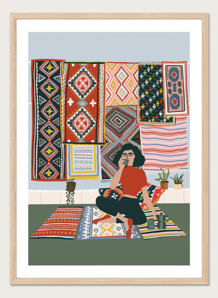 A framed art print featuring a stylized illustration of a woman sitting cross-legged on a patterned rug, surrounded by colorful geometric textiles hanging on a wall. Bohemian Coffee Morning by Ceyda Alasar interior art print featuring patterned rugs, floor cushions, potted plants, coffee, and colorful collected decor.