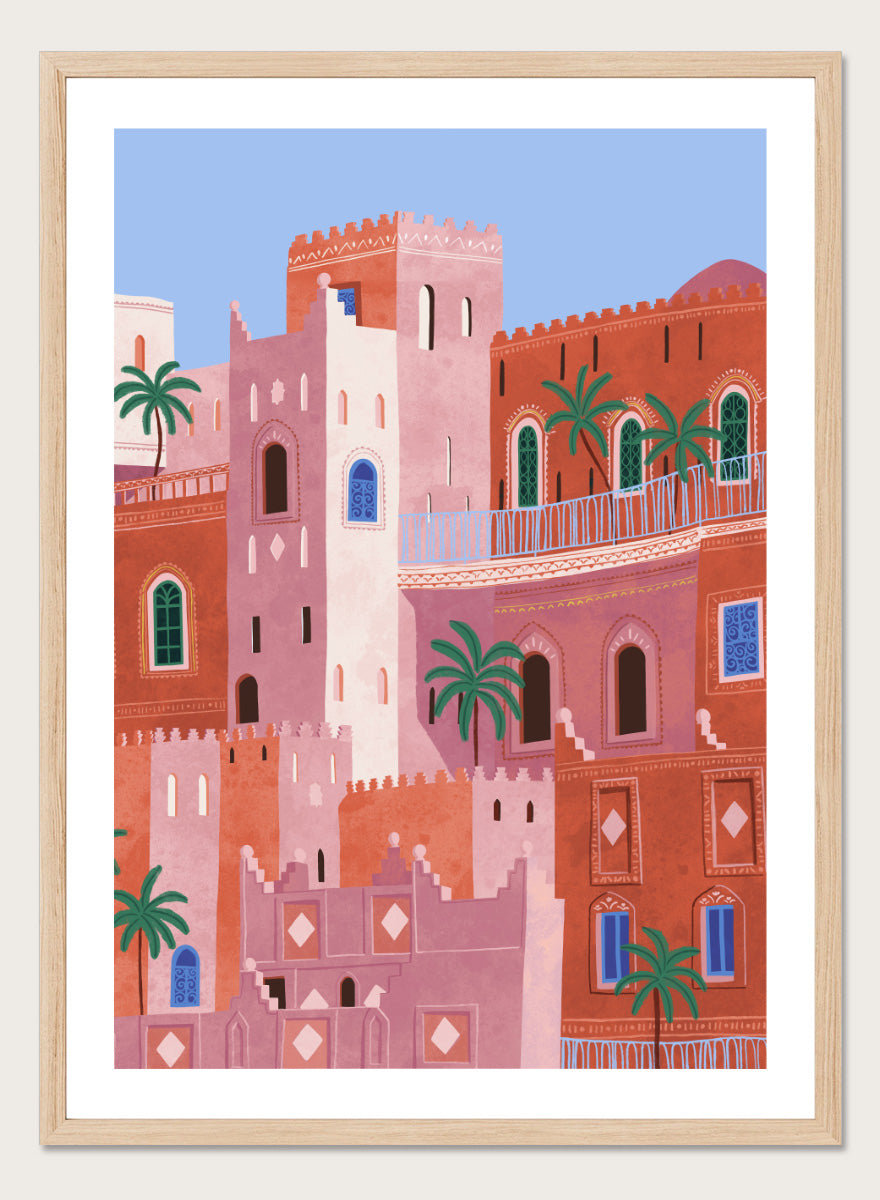 A framed art print featuring a stylized illustration of Moroccan-style buildings in shades of terracotta, pink, and cream, accented with green palm trees and blue window details, set against a light blue sky. Vibrant Moroccan City by Ceyda Alasar travel art print featuring layered terracotta architecture, arched windows, palms, and colorful Moroccan details.