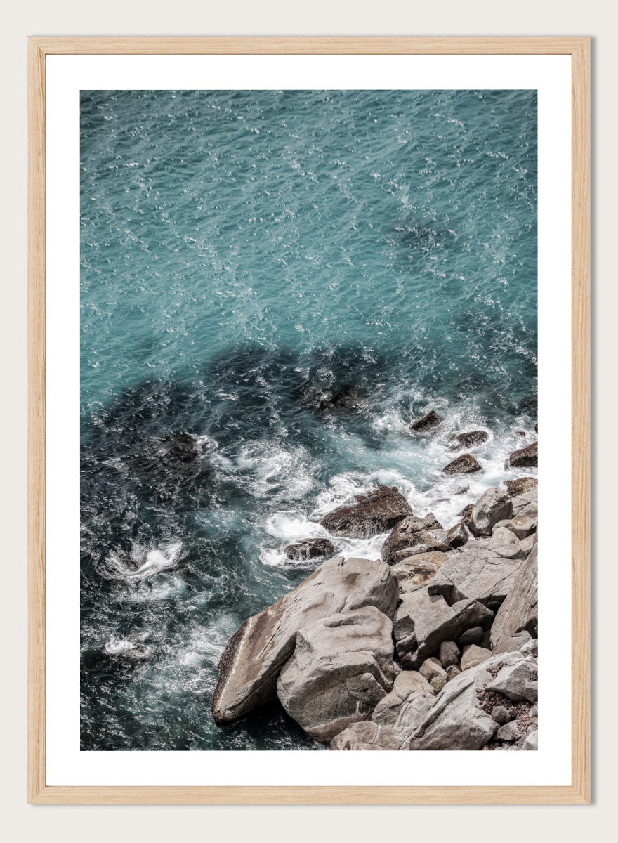 A framed photographic art print depicting a top-down view of turquoise ocean water washing over dark rocks and a rocky shoreline. Ocean’s Edge Symphony by Shot by Clint aerial ocean photography print featuring turquoise water, white surf, coastal texture, and soft seafoam tones.