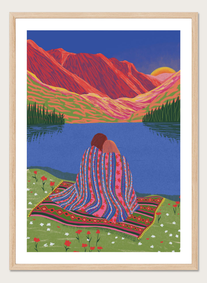 A framed art print featuring a stylized landscape illustration of two people sitting together on a patterned blanket by a blue lake, with red mountains and a yellow sun in the background. Cozy Lake Sunset art print by Ceyda Alasar features a lakeside embrace, vivid mountains, and a glowing sunset in rich, story-filled color.