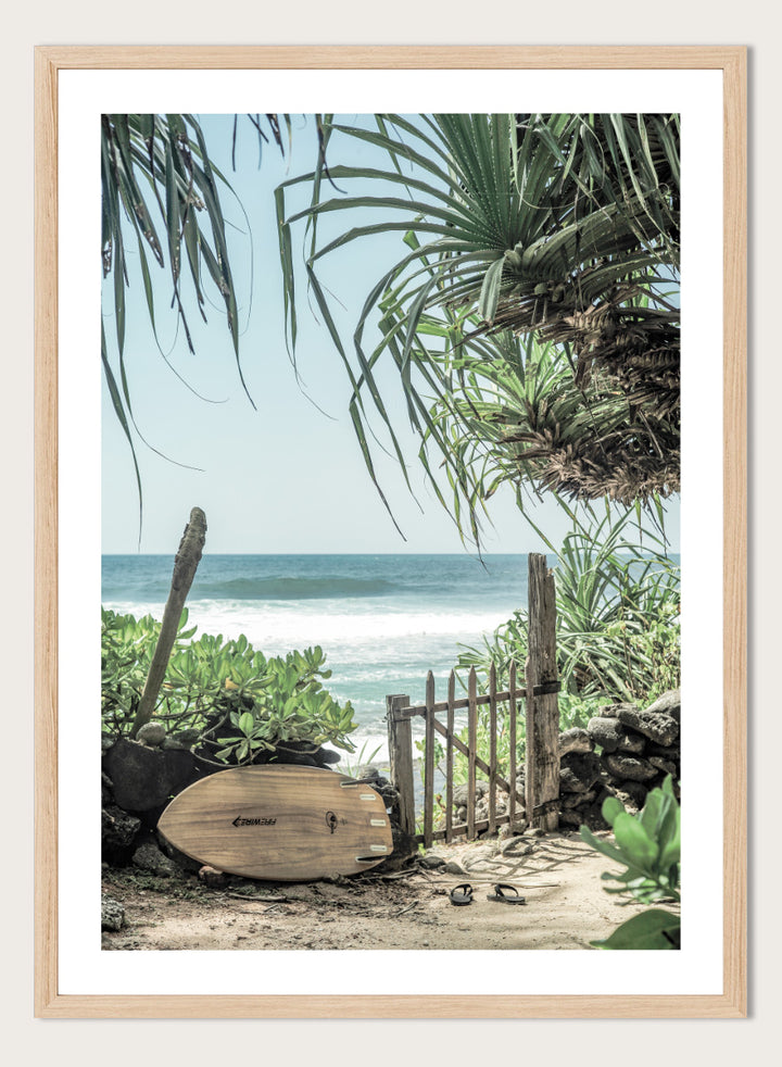 A framed beach scene art print featuring a wooden surfboard leaning against a rustic gate, with palm leaves framing a view of the ocean under a light blue sky. Coastal Serenity by Shot by Clint coastal art print featuring a surfboard, tropical plants, rustic gate, sandy path, and ocean waves.