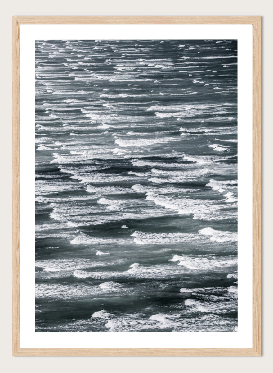 A framed black and white photographic print depicting a close-up view of ocean waves with white foam crests against dark water. Waves in Rhythm by Shot by Clint ocean photography print featuring layered blue-grey waves, white foam, and powerful natural movement across the sea.