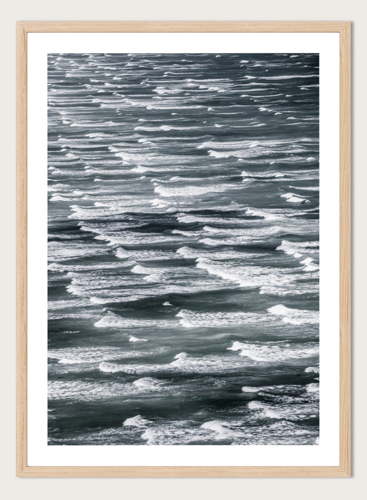 A framed black and white photographic print depicting a close-up view of ocean waves with white foam crests against dark water. Waves in Rhythm by Shot by Clint ocean photography print featuring layered blue-grey waves, white foam, and powerful natural movement across the sea.