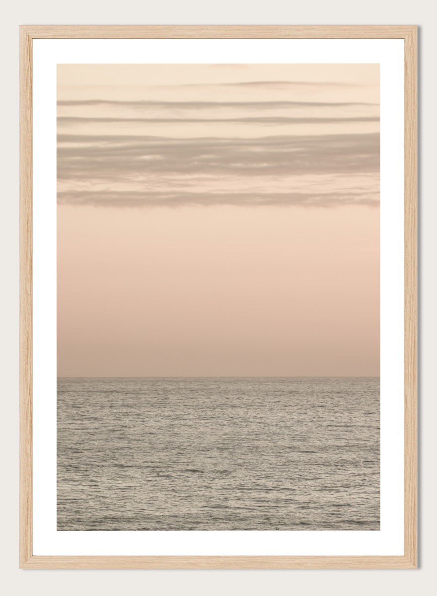 A minimalist framed art print depicting a calm seascape at dusk or dawn, with a soft peach and beige sky meeting a muted gray-blue ocean horizon. Serene Horizons by Shot by Clint features a soft peach sky and calm ocean horizon in a minimal coastal photograph with airy neutral tones.
