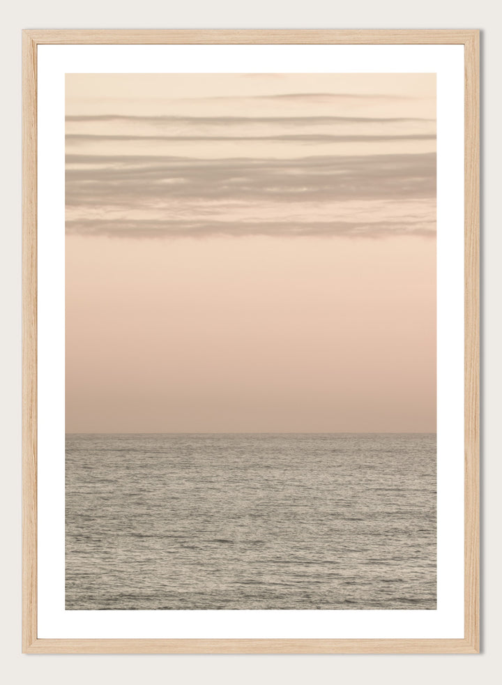 A minimalist framed art print depicting a calm seascape at dusk or dawn, with a soft peach and beige sky meeting a muted gray-blue ocean horizon. Serene Horizons by Shot by Clint features a soft peach sky and calm ocean horizon in a minimal coastal photograph with airy neutral tones.