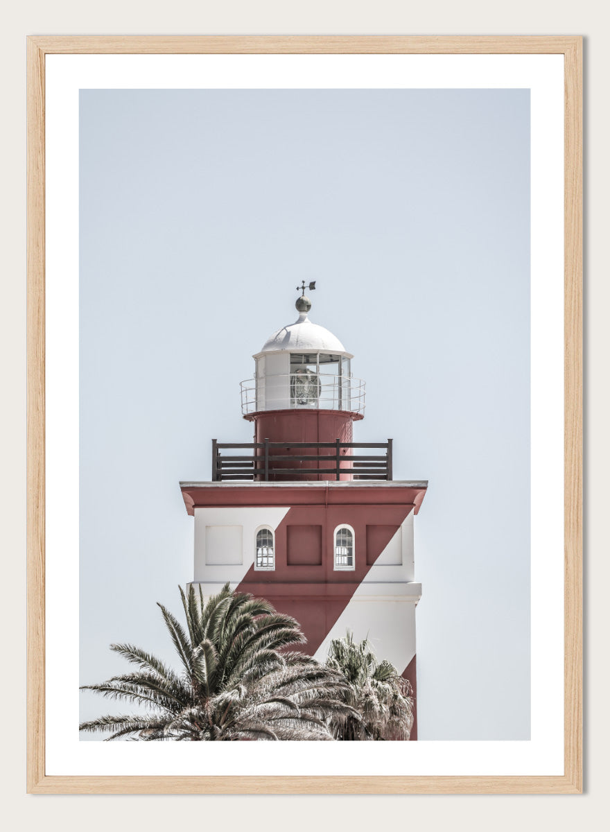 A framed art print featuring a red and white lighthouse with palm trees at the bottom, set against a pale sky background. Beacon of Solitude by Shot by Clint lighthouse photography print featuring red-and-white architecture, palm trees, arched windows, and a pale coastal sky.