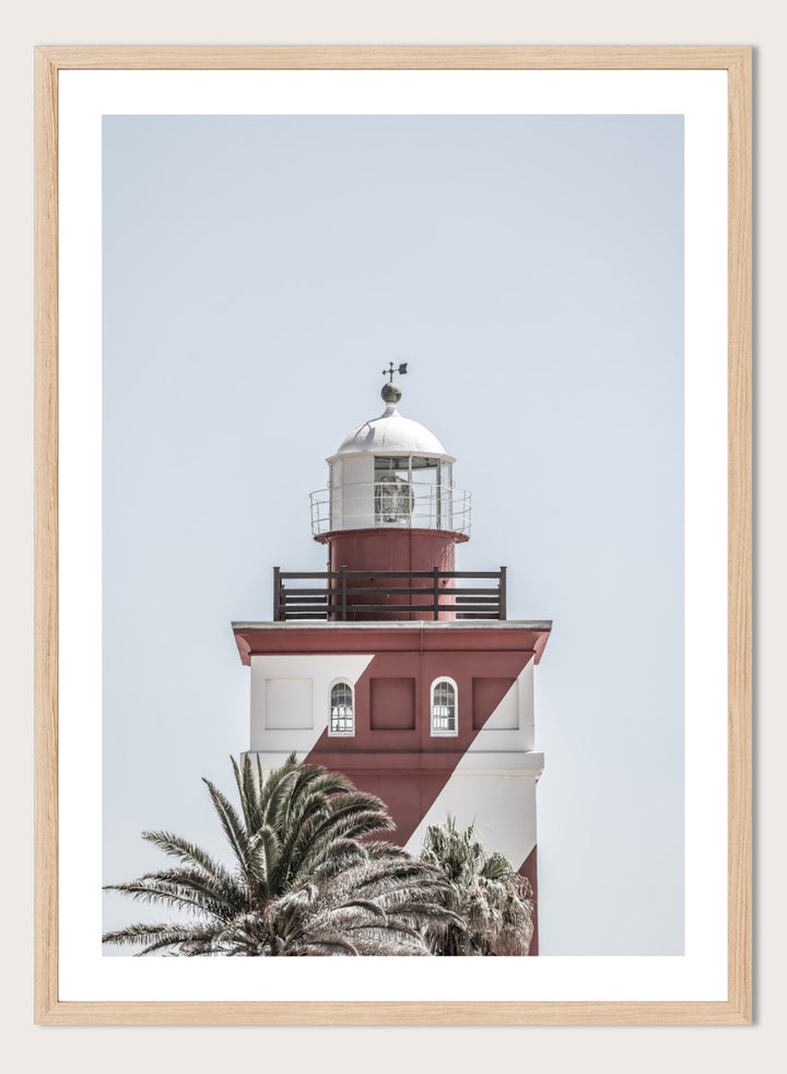 A framed art print featuring a red and white lighthouse with palm trees at the bottom, set against a pale sky background. Beacon of Solitude by Shot by Clint lighthouse photography print featuring red-and-white architecture, palm trees, arched windows, and a pale coastal sky.