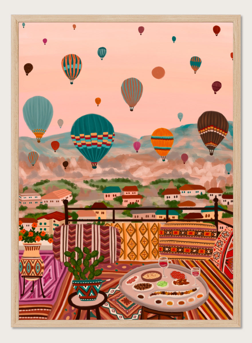 A colorful art print depicting a scenic view of a hillside town with mountains in the background, filled with hot air balloons. In the foreground, a patterned outdoor seating area features a table with food and a potted plant. Shop Balloons Over Rooftop Dinner by Nidhi Kachhadiya, a colorful travel wall art print featuring hot air balloons, rooftop dining, patterned textiles, and teal and coral tones.