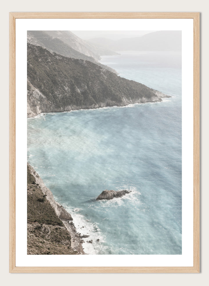 A framed photographic art print depicting a coastal landscape with a steep rocky cliff overlooking a calm, pale blue sea with a small rock formation in the foreground. Coastal Serenity by Shot by Clint coastal photography print featuring pale blue water, rugged cliffs, rocky shores, and a soft hazy horizon.