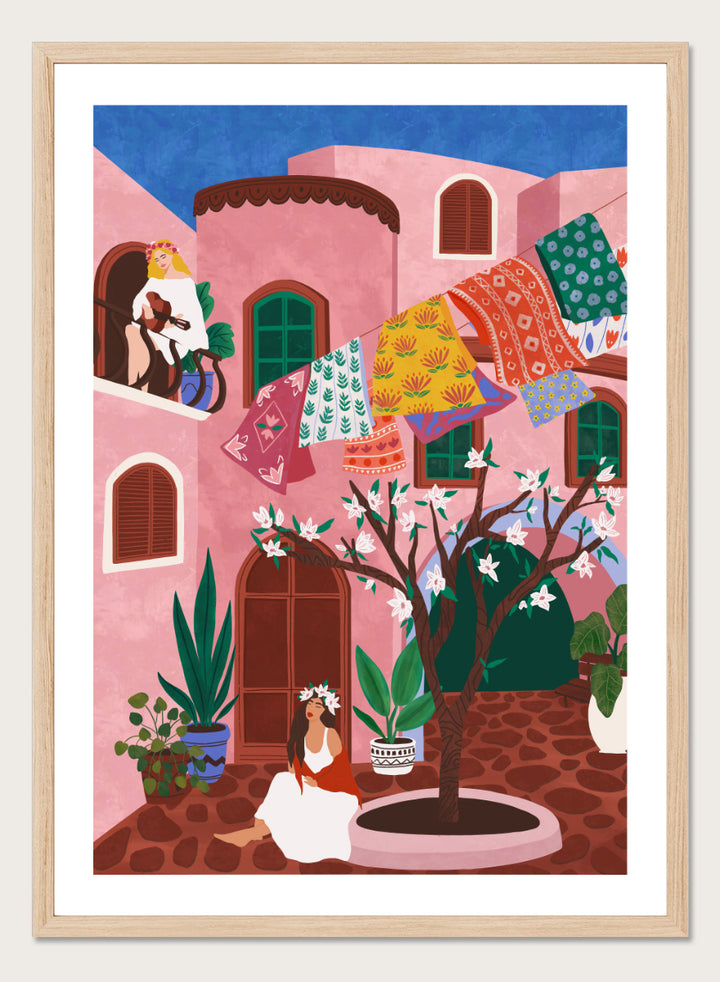 A framed art print featuring a colorful Mediterranean-style illustration of a pink courtyard with a woman sitting under a flowering tree, laundry hanging in the background, and a blue sky above. Pink Village Courtyard by Ceyda Alasar art print featuring pink architecture, patterned rugs, flowering trees, potted plants, and relaxed courtyard life.