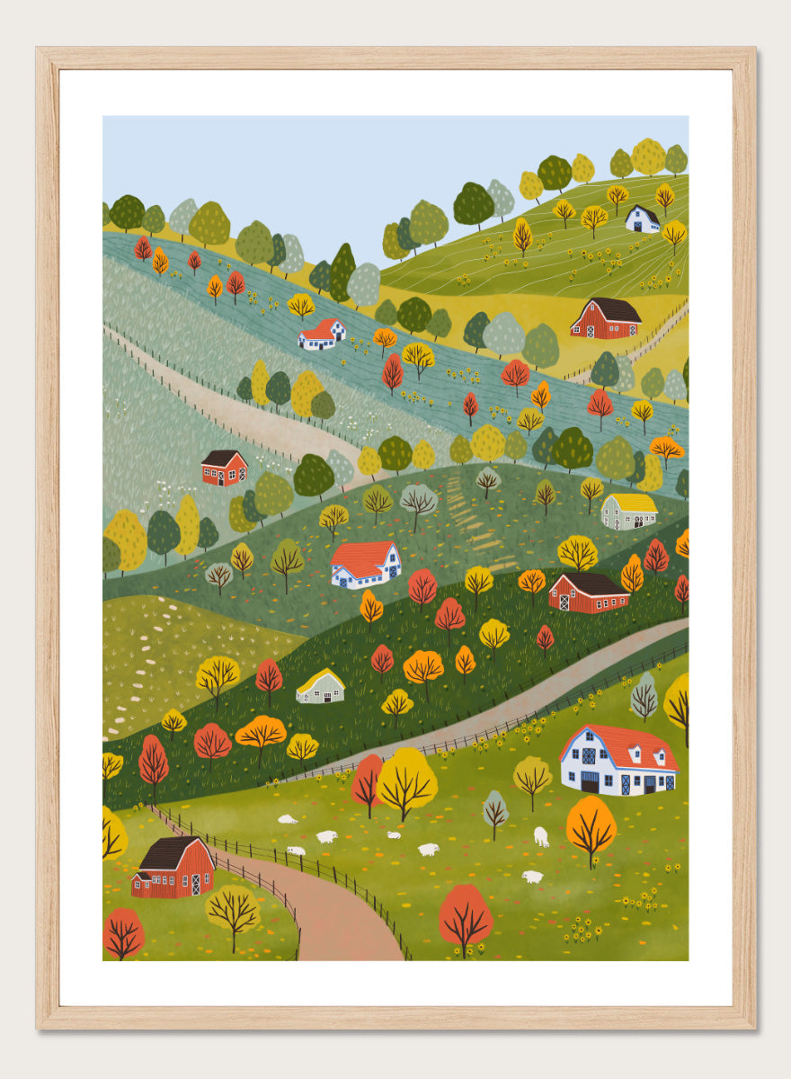 A framed art print featuring a whimsical, stylized illustration of a rolling green and yellow countryside landscape with red houses, colorful autumn trees, winding roads, and a pale blue sky. Vibrant Autumn Village Hills by Ceyda Alasar art print with rolling countryside, colorful fall trees, farmhouses, barns, sheep, and winding roads.