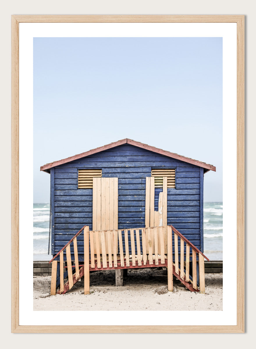 A framed photographic print of a blue wooden beach lifeguard hut on a sandy shore with the ocean and pale sky in the background. Beachside Retreat by Shot by Clint features a weathered blue beach hut and soft ocean backdrop in a calm coastal photography print.
