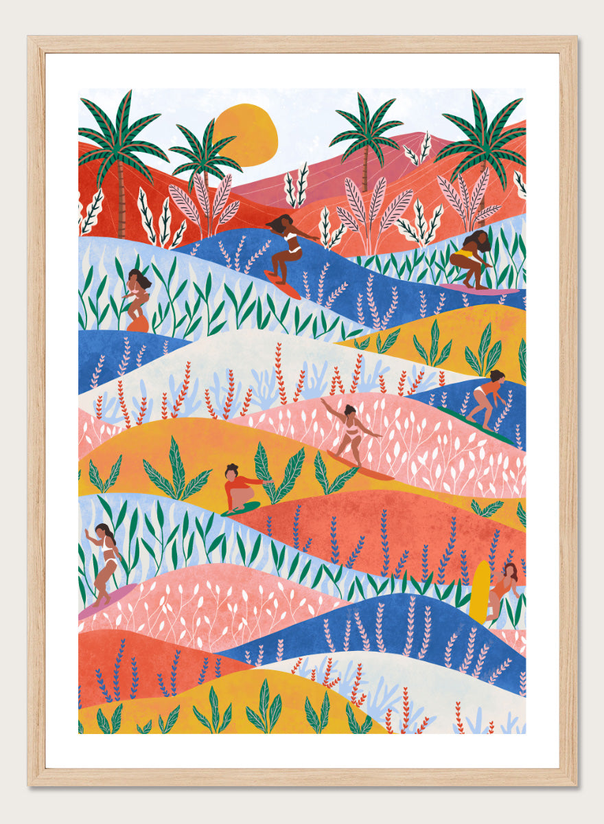 A framed art print featuring a colorful, stylized landscape illustration with rolling hills, palm trees, a sun, and small human figures in a folk-art style. Tropical Surf Girls by Ceyda Alasar surf art print featuring women riding waves through a colorful palm-filled landscape in bold tropical tones.