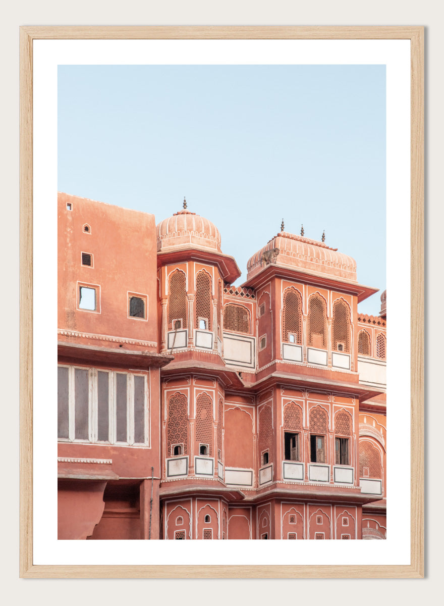 A framed art print featuring a photograph of the iconic pink sandstone Rajasthani palace, Udaipur, with white architectural details under a clear light blue sky. Terracotta Splendor by Shot by Clint architectural photography print featuring ornate palace windows, domed balconies, carved details, and warm coral tones.