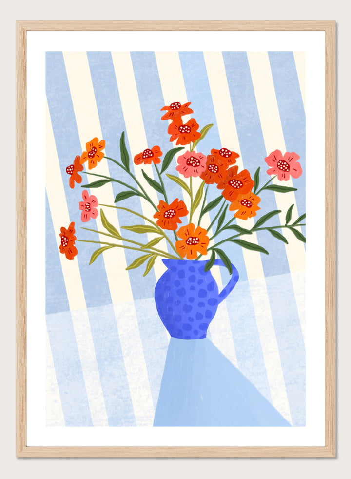 A framed art print featuring a stylized illustration of a blue patterned pitcher vase filled with orange, red, and pink flowers against a blue and white striped background. Cheerful Floral Pitcher by Bea Müller floral still life art print featuring orange and pink flowers in a bright blue jug with striped blue decor.