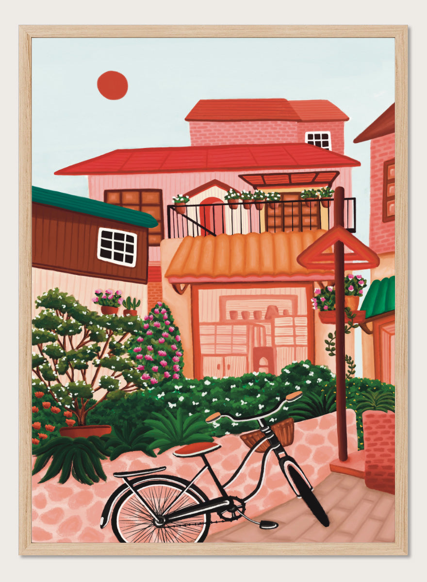 A colorful illustrated street scene with pink and peach buildings, green plants, flowers, a bicycle in the foreground, and a pale sky with a red sun. Shop Charming Village Street by Nidhi Kachhadiya, a colorful village wall art print featuring a bicycle, flower-filled homes, terracotta roofs, and warm pink and green tones.