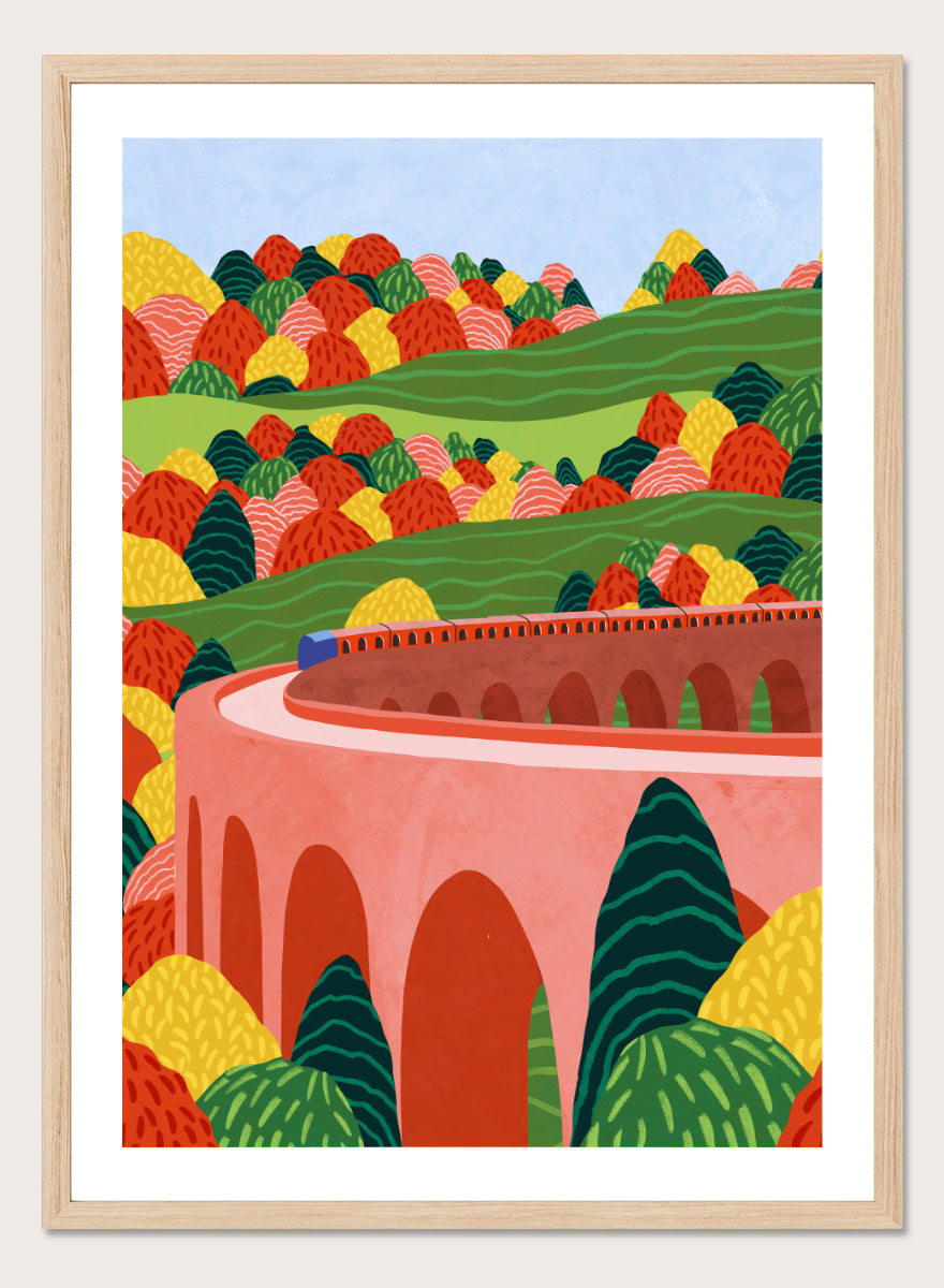 A framed art print featuring a stylized landscape illustration with rolling green hills, colorful patterned trees, and a pink arch bridge under a light blue sky. Winding Autumn Viaduct by Ceyda Alasar travel art print featuring a colorful train, arched bridge, rolling hills, and bold autumn foliage.