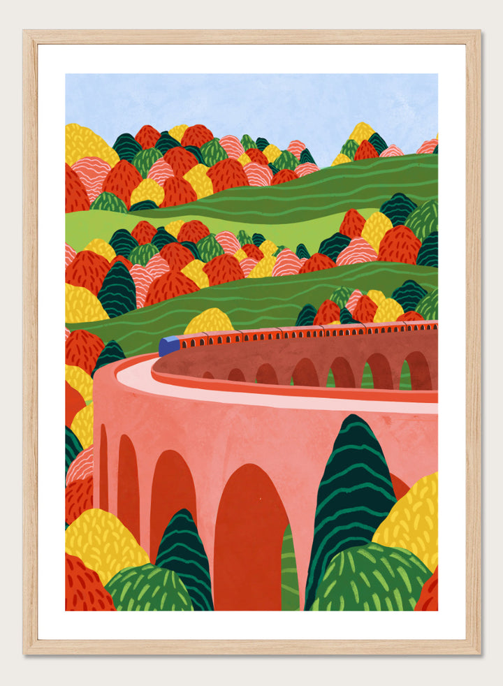 A framed art print featuring a stylized landscape illustration with rolling green hills, colorful patterned trees, and a pink arch bridge under a light blue sky. Winding Autumn Viaduct by Ceyda Alasar travel art print featuring a colorful train, arched bridge, rolling hills, and bold autumn foliage.