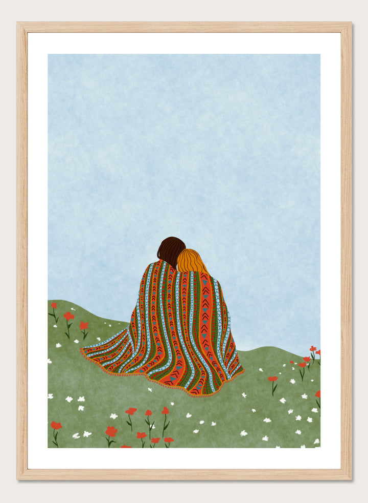 A framed art print featuring an illustration of two people sitting on a grassy hill, wrapped in a vibrant, patterned blanket under a light blue sky. Cozy Hillside Embrace by Ceyda Alasar art print featuring two people wrapped in a patterned blanket among green hills and wildflowers.