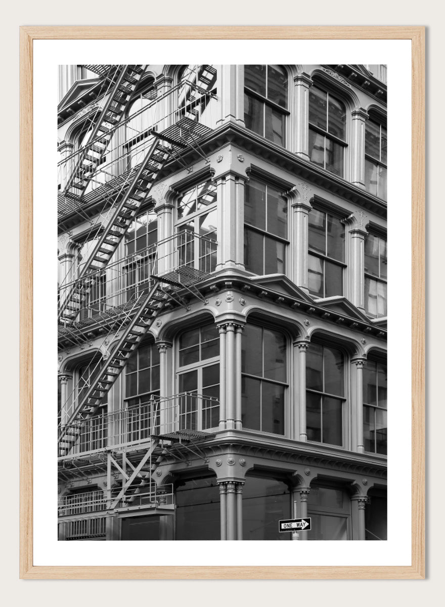 Black and white architectural photograph of a corner building facade with ornate windows and external fire escapes. Urban Geometry by Shot by Clint black-and-white architectural photography print featuring fire escapes, tall windows, ornate columns, and layered city detail.