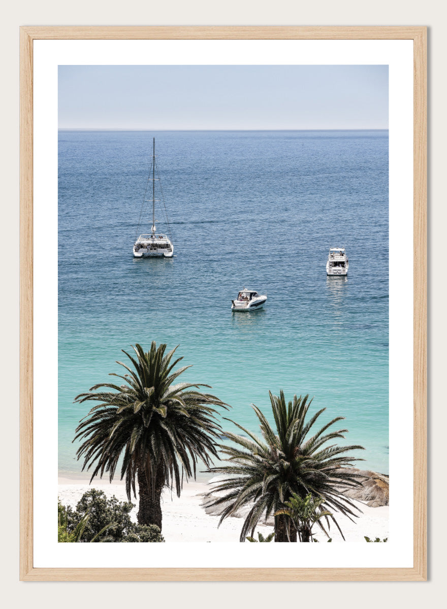 A framed coastal photograph showing a calm blue ocean with three boats, turquoise water near a white sandy shore, and two palm trees in the foreground. Tropical Serenity by Shot by Clint coastal photography print featuring turquoise water, sailboats, palm trees, and a bright tropical shoreline.