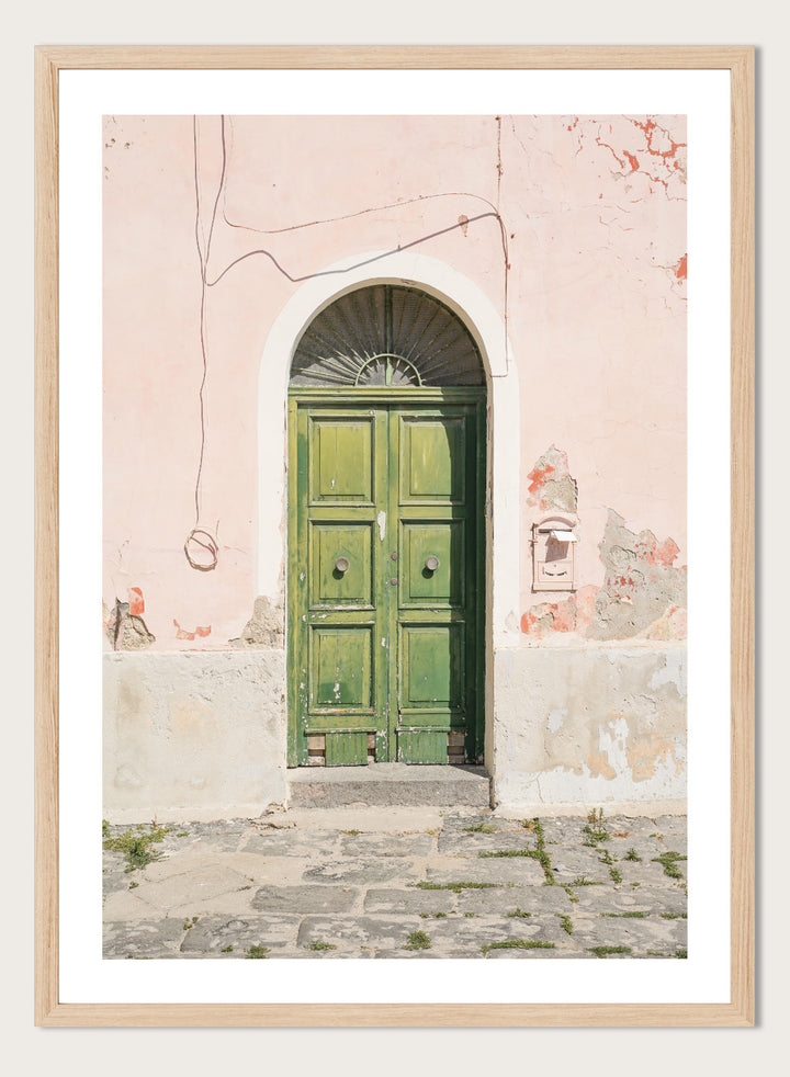 Green door on a pink wall with peeling paint. Old World Green Door by Henrike Schenk features a vintage European doorway, weathered architecture, pastel walls, travel photography, and wall art.