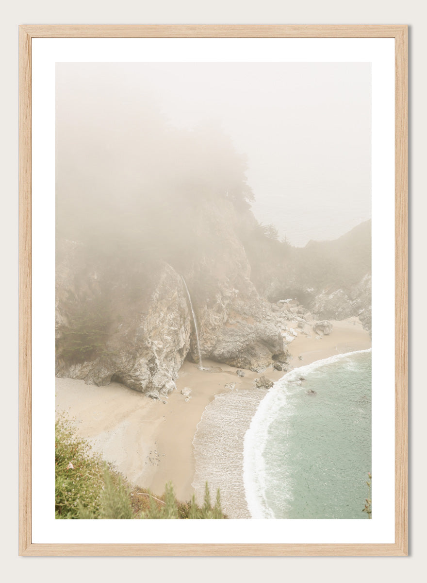 A framed photographic print of a misty coastal beach scene with a small waterfall, sandy shore, and pale blue-green ocean waves. Misty Coastal Haven by Shot by Clint coastal photography print featuring a secluded cove, rocky cliffs, pale turquoise water, and a soft veil of mist.