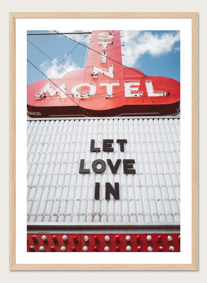 Vintage motel sign with 'Let Love In' message against a blue sky. Let Love In by Bethany Young features a vintage motel sign with red neon lettering against blue sky. Retro Americana photography wall art print.