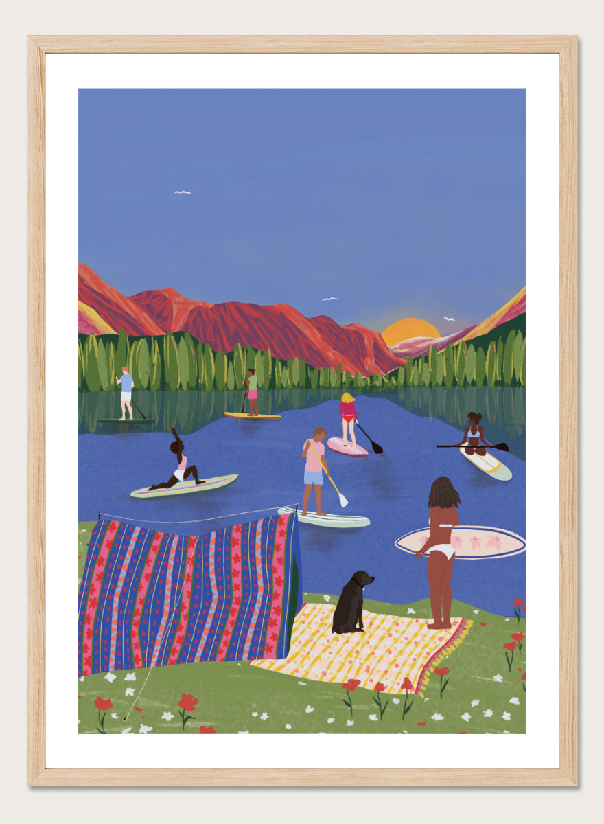 A framed art print featuring a colorful, stylized illustration of people paddleboarding and relaxing on a lake with mountains in the background. Paddleboard Lake Life by Ceyda Alasar outdoor art print featuring paddleboarders, mountain scenery, wildflowers, picnic blankets, and a lakeside dog.