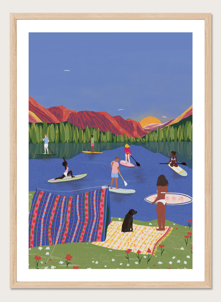 A framed art print featuring a colorful, stylized illustration of people paddleboarding and relaxing on a lake with mountains in the background. Paddleboard Lake Life by Ceyda Alasar outdoor art print featuring paddleboarders, mountain scenery, wildflowers, picnic blankets, and a lakeside dog.