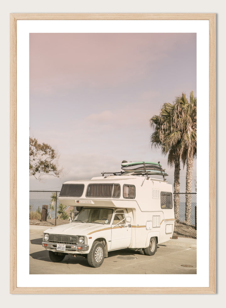 A vintage white camper truck parked outdoors near palm trees and a fence, with a surfboard strapped to its roof rack. Vintage Camper Parked by Shot by Clint travel photography print featuring a retro camper, surfboards, palm trees, and sun-faded coastal tones.
