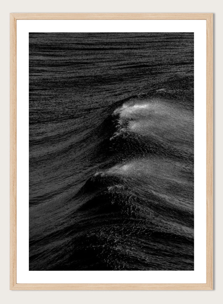 Black and white fine art photographic print of ocean waves, featuring a dramatic wave crest with white foam against dark textured water. Night Wave Cascade by Shot by Clint black-and-white ocean photography print featuring dark rolling waves, sea spray, and dramatic natural movement.