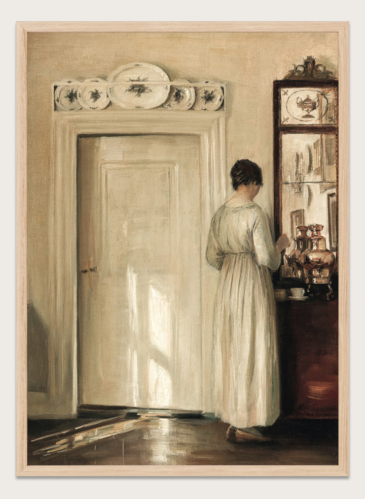 Woman in a white dress standing in a room with a door and shelves filled with items. Woman Making Tea by Carl Holsøe is a vintage Danish interior art print featuring Scandinavian home interiors, muted tones, natural light, and a woman preparing tea.