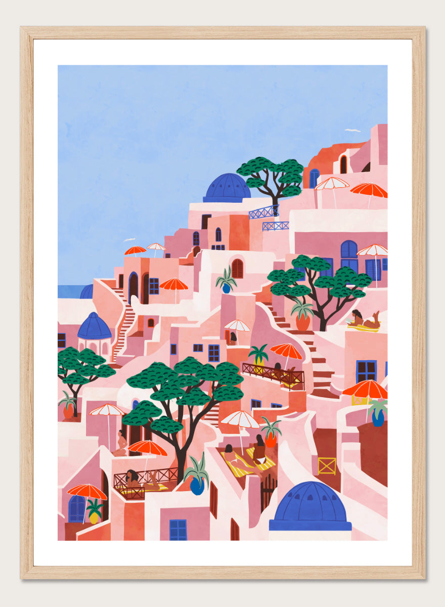 A stylized art print depicting a hillside Mediterranean village with pink and peach buildings, blue domes, green trees, and a light blue sky. Santorini Summer Dream by Ceyda Alasar art print featuring blue domes, terraced Mediterranean architecture, orange umbrellas, and sunny Greek island charm.
