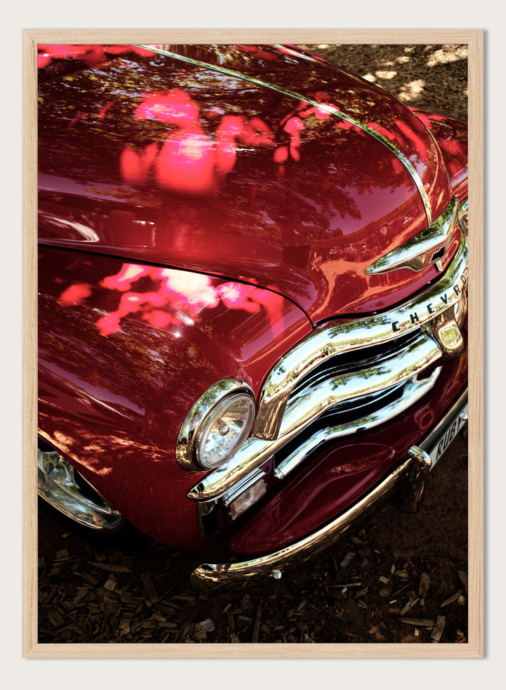 Close-up of a vintage red Chevrolet car body with chrome grille and reflections of trees on the glossy paint, photographed outdoors on mulch. Vintage Red Car by Shot by Clint automotive photography print featuring a classic Chevrolet, deep red paint, polished chrome, and sculptural vintage details.