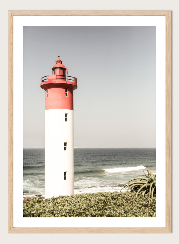 A framed photographic art print of a red and white lighthouse standing on a grassy shore with the ocean and sky in the background. Beacon by the Sea by Shot by Clint coastal photography print featuring a red-and-white lighthouse, ocean waves, pale sky, and coastal greenery.