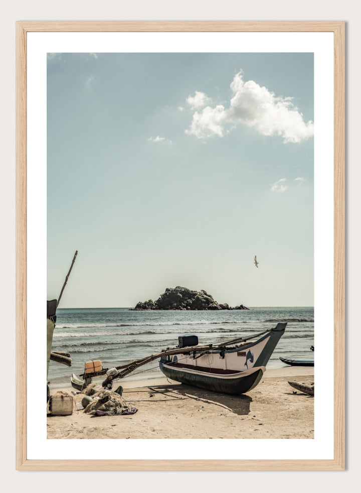 A framed photographic print of a coastal beach scene with a small boat on the sandy shore, the sea and distant rocky island, and a pale blue sky with clouds. Seaside Serenity by Shot by Clint coastal photography print featuring a fishing boat, sandy shore, ocean waves, distant island, and weathered seaside details.