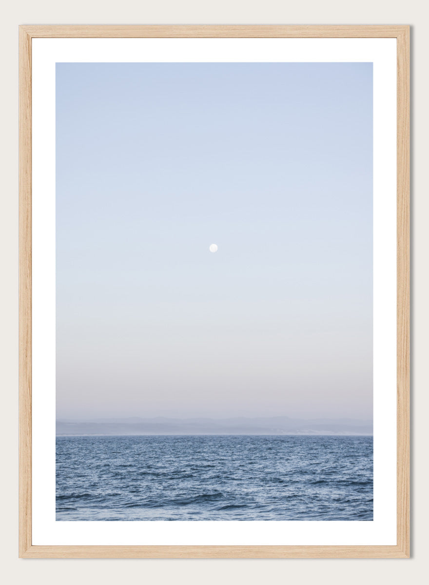 A minimalist framed art print depicting a calm blue ocean under a pale sky with a small visible moon. Moonlit Horizon by Shot by Clint coastal photography print featuring a pale moon, distant shoreline, open blue water, and soft lavender evening tones.