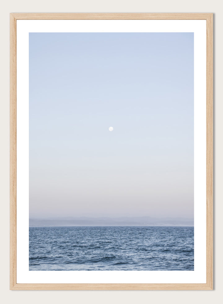 A minimalist framed art print depicting a calm blue ocean under a pale sky with a small visible moon. Moonlit Horizon by Shot by Clint coastal photography print featuring a pale moon, distant shoreline, open blue water, and soft lavender evening tones.