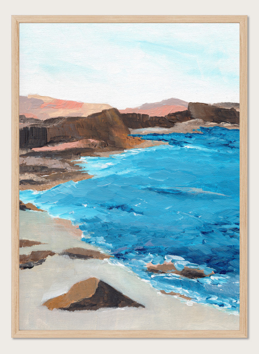 A framed landscape painting depicting a coastal scene with bright blue ocean water, sandy shore, dark rocky cliffs, and distant hills under a pale sky. Shop Rocky Coastal Waters by Nikita Jariwala, a coastal wall art print poster with blue ocean water, rocky cliffs, and a peaceful seaside view.