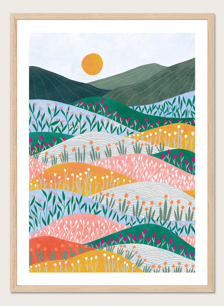 A framed art print featuring a stylized landscape illustration with rolling hills, wildflowers, mountains, and a sun in pastel and bright colors. Colorful Flower Hills by Ceyda Alasar botanical landscape art print featuring layered wildflower fields, rolling hills, mountains, and a golden sun.