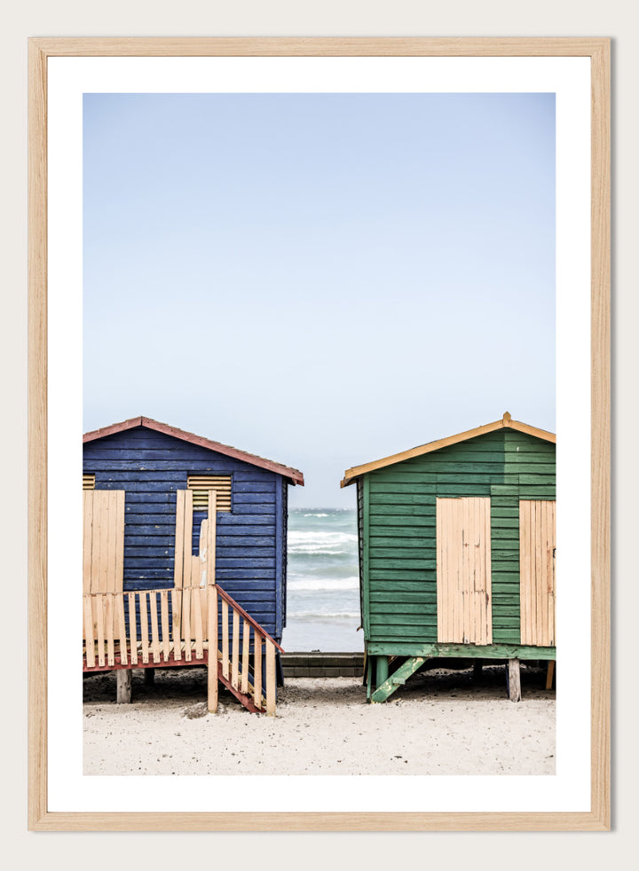 A framed photographic print of two wooden beach huts on a sandy beach under a pale blue sky, with one hut in dark blue and the other in green, both with light wood shutters and doors. Coastal Huts Harmony by Shot by Clint coastal photography print featuring blue and green beach huts, sandy ground, weathered wood, and ocean views.