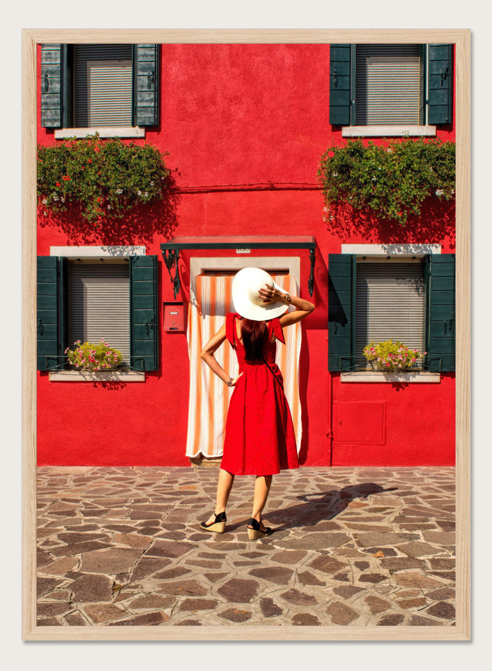 Woman in a red dress and white hat standing in front of a red wall with green shutters. Red Art Print by Anette Ohlendorf featuring a woman in a red dress standing against a red building. Officially licensed travel photography with strong color and symmetry.