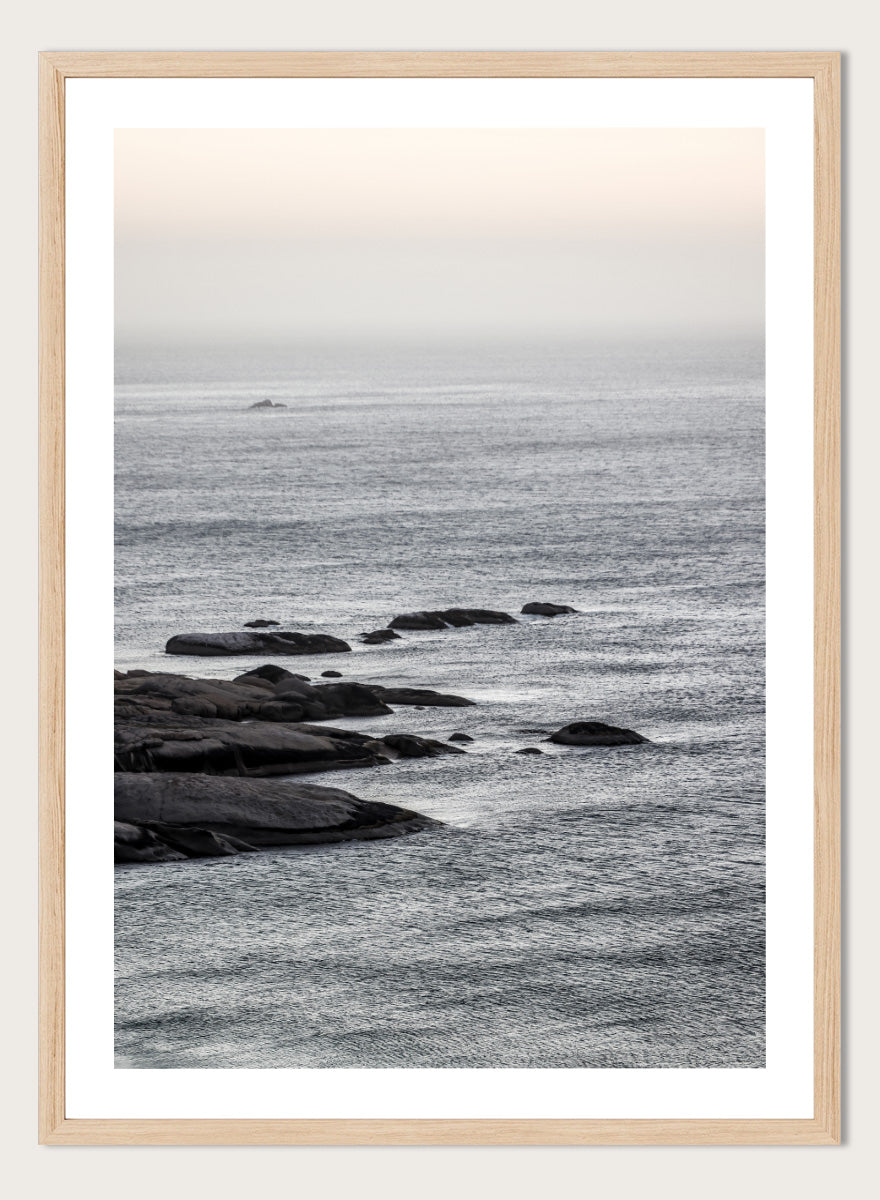 A framed black and white coastal landscape photograph featuring a calm sea, distant rocks, and a pale sky. Silver Sands by Shot by Clint coastal photography print featuring silver-blue water, dark shoreline rocks, and a soft misty horizon.