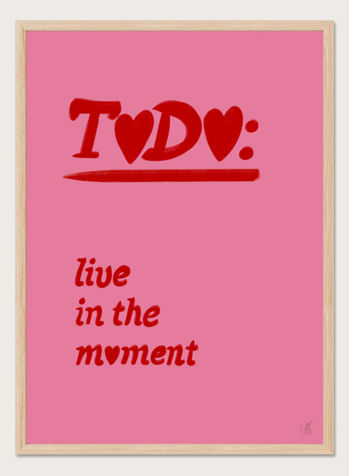 Pink poster with red text 'TODO: live in the moment' on a pink background. Typography wall art reading “live in the moment” with red hearts on pink background by Anne-Marie Volfova. Officially licensed motivational art print.