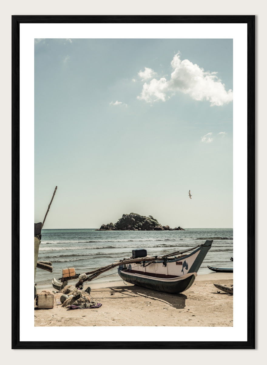 A framed photographic print of a coastal beach scene with a small boat on the sandy shore, the sea and distant rocky island, and a pale blue sky with clouds. Seaside Serenity by Shot by Clint coastal photography print featuring a fishing boat, sandy shore, ocean waves, distant island, and weathered seaside details.